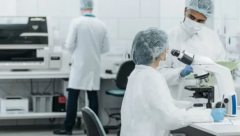Three scientists working in a lab in conversation while one looks through a microscope